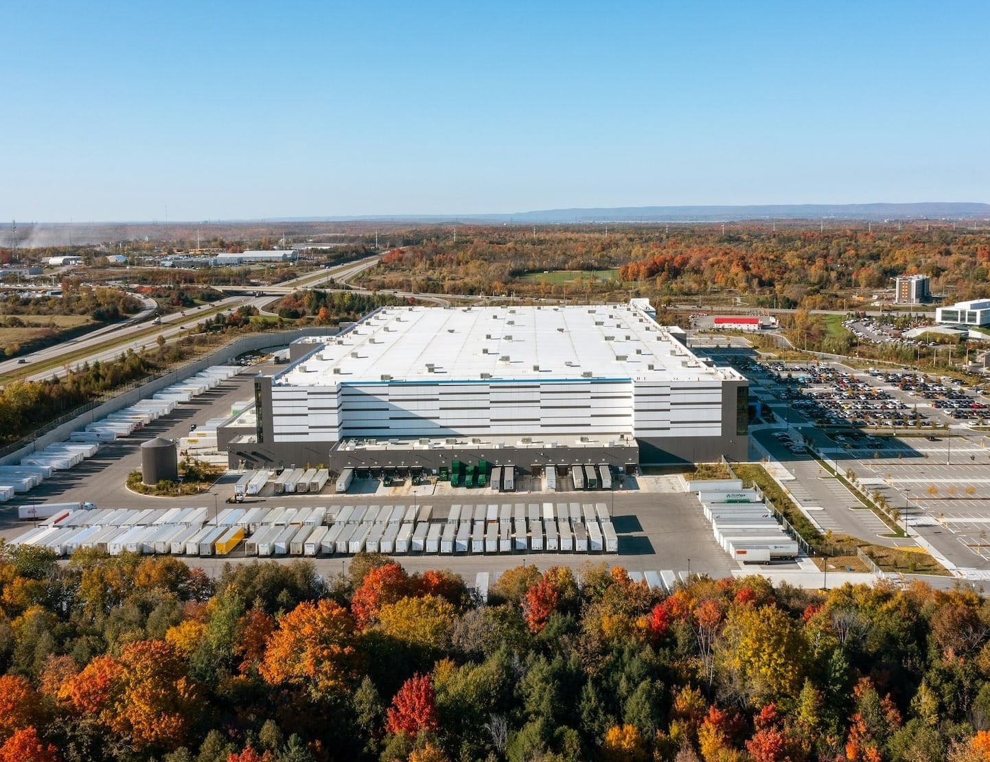 Stort lagerbyggeri med parkeringspladser og lastbiler, omgivet af efterårsskov og veje. Klar himmel.